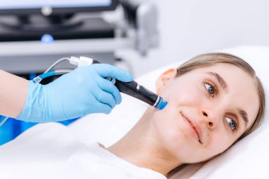 Kadın kozmetikçi, genç bir kadın üzerinde, modern ekipmanlar parlak bir tedavi odasında görülebilirken, bir yüz aygıtı kullanarak hidrakiyal bir prosedür uyguluyor.