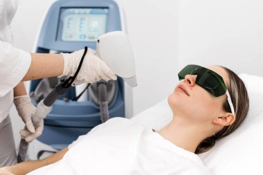 Kadın hasta klinik bir ortamda lazer kılı temizleme tedavisi görüyor. Teknisyen olarak koruyucu gözlük takıyor ve prosedür için lazer cihazı kullanıyor.