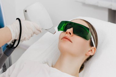 Koruyucu gözlüklü bir kadın hasta, temiz bir klinik ortamında tedavi yatağında yatarken yüzünde lazer tüyü alınıyor.
