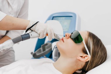 Klinik ortamda koruyucu gözlüklerle lazer temizleme tedavisi gören kadın gelişmiş makineler ve arka planda görünen aletler.