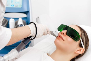 Çağdaş bir klinikte lazer tedavisi gören bir kadın koruyucu gözlük takıyor ve tıbbi malzemelerle tedavi yatağında yatıyor.