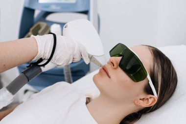 Klinik ortamda lazer tedavisi gören bir kadın koruyucu gözlük takıyor ve yanında tıbbi aletlerle tedavi yatağında yatıyor.