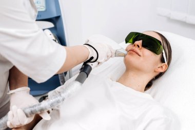 Klinik ortamda lazer tedavisi gören bir kadın koruyucu gözlük takıyor ve cildinde lazer cihazı kullanıyor.