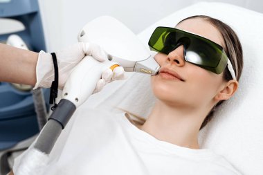 Klinik ortamda lazer tedavisi gören bir kadın koruyucu gözlük takıyor. Bir teknisyen ise lazer cihazını parlak bir ayarda çalıştırıyor.