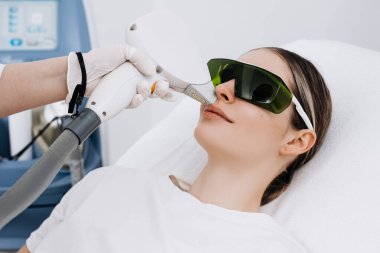 Kadın hasta klinik bir ortamda lazer kılı temizleme tedavisi görüyor koruyucu gözlük takıyor ve yanında ekipmanlarıyla tedavi yatağında yatıyor.