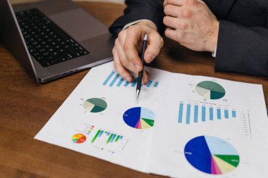 Takım elbiseli bir işadamı, arka planda dizüstü bilgisayarı ve ahşap bir masada çeşitli grafik ve çizelgeleri içeren mali belgeleri inceliyor.