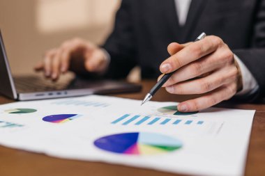 İşadamı, veri analizi için yakınlarda bir dizüstü bilgisayarla birlikte bir masanın üzerindeki grafik ve çizelgeleri inceleyen mali belgeleri inceliyor.
