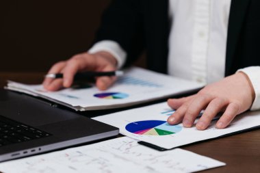 İş adamı, ofis ortamındaki ahşap bir masada dizüstü bilgisayarın yanında turta grafikleri ve grafiklerin yer aldığı mali belgeleri inceliyor.