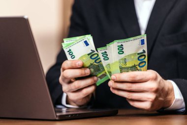 Siyah takım elbiseli bir adam, bir masa başında dizüstü bilgisayarla 100 Euro 'luk banknotlar sayıyor. Ofis ortamında mali yönetim ve para yönetimini gösteriyor.