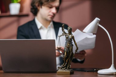 Erkek avukat, masa başında dizüstü bilgisayarı ve bayan adalet heykeliyle birlikte, sıcak tonlu çağdaş ofis ortamını inceliyor.