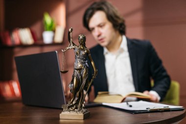 Erkek avukat masa başında otururken dizüstü bilgisayara odaklanıyor, ön planda Adalet Heykeli ve hukuk kitapları ile modern ofis dekorasyonu olay yerini büyütüyor