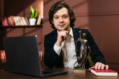 Siyah takım elbiseli bir erkek avukat elinde dizüstü bilgisayar, hukuk kitapları ve kadın adalet heykeliyle bir masada oturup profesyonel bir ofis ortamı yaratıyor.