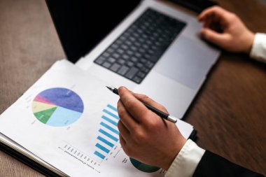 İşadamı, ofis ortamında ahşap bir masada dizüstü bilgisayar kullanırken finansal verileri pasta grafiği ve bar grafiğiyle inceliyor.