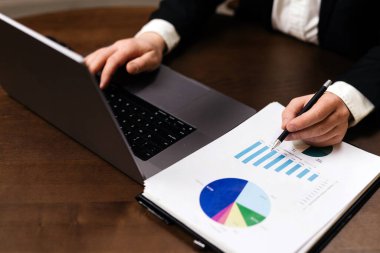 İş adamı, veri analizi ve sunum için bir dizüstü bilgisayar kullanarak ahşap bir masadaki tablolar ve grafiklerle mali belgeleri gözden geçiriyor.