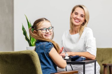 Gözlüklü genç bir kız beyaz elbiseli bir kadınla aynı masada oturuyor. İkisi de kapalı bir ortamda gülümsüyor. Yakınlarda bir saksı bitkisi var.