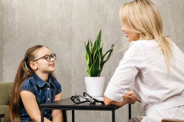 Çağdaş ofis ortamında gözlük takan genç bir kız, masanın üzerinde saksı ve tıbbi aletler bulunan bir kadın doktorla etkileşime giriyor.
