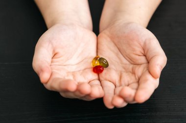 Çocuğun elleri koyu ahşap bir yüzey üzerinde kırmızı, sarı ve yeşil renkli üç vitamin vitamini sergiler, canlı şekilleri ve dokuları vurgular.