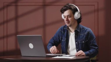 Kulaklıklı genç adam ahşap bir masada otururken dizüstü bilgisayarına odaklanıyor.