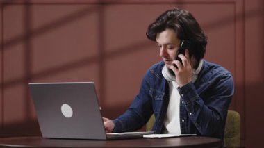 Kulaklıklı genç adam ahşap bir masada otururken dizüstü bilgisayarına odaklanıyor.