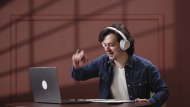 Kulaklıklı genç adam ahşap bir masada otururken dizüstü bilgisayarına odaklanıyor.