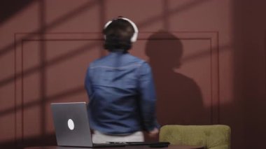 Kulaklıklı genç adam ahşap bir masada otururken dizüstü bilgisayarına odaklanıyor.