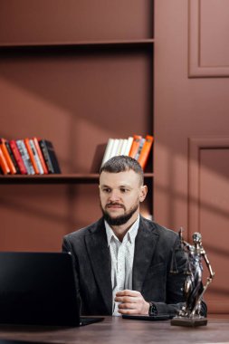 Takım elbiseli bir erkek avukat dizüstü bilgisayarlı bir masada çalışıyor. Kadın Adalet Heykeli ve arka planda renkli kitaplarla dolu kitaplıklar var.