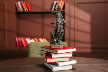 Ahşap bir masanın üzerindeki kitap yığınına yerleştirilmiş Adalet Heykeli. Bulanık bir kitaplık ve yumuşak gölgeler ılık bir atmosfer yaratıyor.