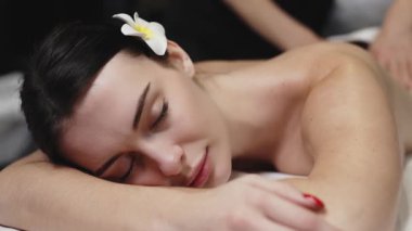Karın üstü uzanan bir kadın masaj masasına sırt masajı yaptırıyor. Etrafı yumuşak ışıklandırma ve rahatlatıcı renklerle dolu sakin bir spa setiyle çevrili.