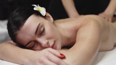 Karın üstü uzanan bir kadın masaj masasına sırt masajı yaptırıyor. Etrafı yumuşak ışıklandırma ve rahatlatıcı renklerle dolu sakin bir spa setiyle çevrili.