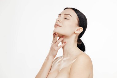 Pürüzsüz ciltli ve doğal güzelliği olan bir kadın, çene üzerinde elleriyle poz vererek, parlak beyaz bir arka planda yüz hatlarını ön plana çıkarıyor.