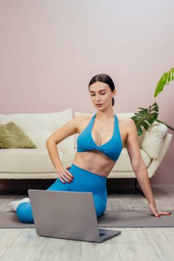 Mavi spor elbiseli bir kadın şık bir oturma odasında yoga yapıyor, dizüstü bilgisayarı ve beyaz bir kanepenin yanında yeşil bir bitkiyle.