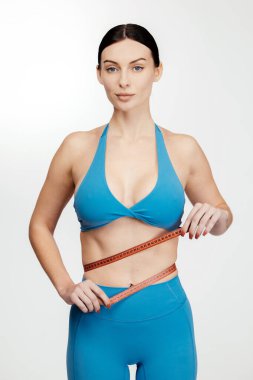 Mavi spor giysili bir kadın, beyaz bir arka planın önünde duran ve vücut şeklini ve zindeliğini vurgulayan bir mezura ile belini ölçer.