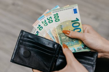 Ellerinde farklı değerlerde Avro banknotlarıyla dolu, hafif ahşap bir yüzeye dayanan siyah bir cüzdan tutarken görülüyor.