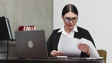 Ofis masasında dizüstü bilgisayarla oturmuş evrak okuyan beyaz bir kadın.