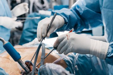 Mavi önlük ve eldiven giyen bir kalp cerrahı ameliyathanede ameliyat aletleri kullanıyor ve arka planda tıbbi ekipmanlar var.