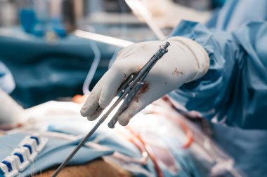 Mavi önlük ve eldiven giyen bir kalp cerrahı ameliyat masasında ameliyat yaparken ameliyat aletleri tutuyor.