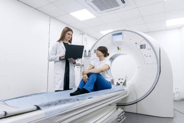 Kadın radyolog muayene masasında hastayla iletişim kuruyor. Parlak, modern bir hastanenin radyoloji bölümünde CT taramasına hazırlanıyor.