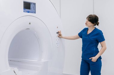 Mavi önlüklü kadın teknisyen, temiz beyaz duvarlar ve gelişmiş görüntüleme ekipmanlarıyla modern bir tıbbi tesiste MRI cihazının ayarlarını ayarlıyor.