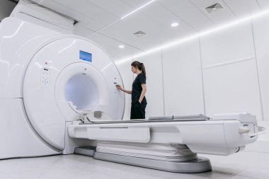 Kadın teknisyen, gelişmiş görüntüleme teknolojisi ve steril bir atmosfer sunarak MRI cihazının ayarlarını çağdaş bir tıbbi ortamda ayarlıyor.
