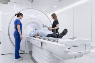 Hasta MRI makinesinde yalan söylerken, iki sağlık uzmanı prosedürü temiz ve iyi aydınlatılmış bir tıbbi muayene odasında izleyip yardım ediyorlar.