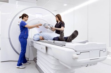 MRI makinesine yerleştirilmiş hasta, önlüklü iki kadın çağdaş bir tıbbi ortamda tarama sürecine yardım ediyor.