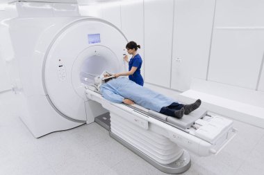 Teknisyen olarak MRI makinesine yerleştirilmiş hasta, parlak ışıklandırma ve steril çevresi olan modern bir tıbbi tesiste taramaya hazırlanıyor.