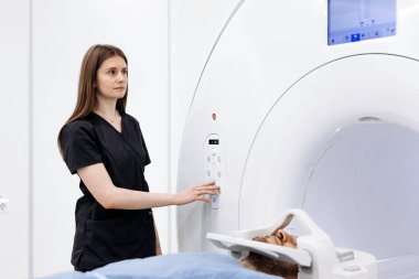 Tıbbi teknisyen MRI cihazındaki ayarları ayarlıyor. Hasta muayene masasında iyi aydınlatılmış, modern bir tıbbi ortamda yatıyor.