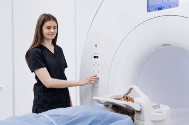 Siyah önlüklü kadın teknisyen, hasta muayene masasında dinlenirken tıbbi tesisteki mavi çarşafla kaplı MRI cihazını ayarlıyor.