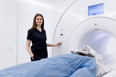 Siyah önlüklü kadın teknisyen MRI makinesi ayarlarını ayarlarken, hasta modern bir tıbbi ortamda mavi çarşafla kaplı muayene masasında yatıyor.