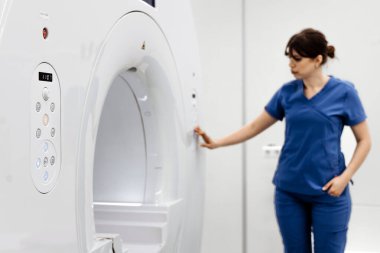 Mavi önlüklü kadın teknisyen, modern bir tıbbi tesiste MRI cihazıyla etkileşime geçiyor. Gelişmiş görüntüleme teknolojisi ve klinik ayarlar sergiliyor.