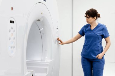 Mavi önlüklü kadın teknisyen, modern bir tıbbi tesiste MRI cihazının ayarlarını düzelterek, temiz ve iyi aydınlatılmış bir sağlık ortamını gösteriyor.