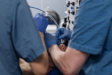 Tıbbi personel klinik ortamda mavi önlük ve eldiven giyen ve arka planda ekipmanı olan bir hastaya endotrakeal entübasyon uygular.