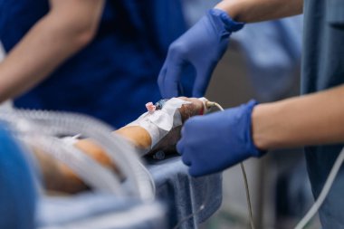 Mavi önlüklü tıbbi personel, arka planda tıbbi ekipman ve klinik ortam bulunan hastanın koluna serum bağlar.