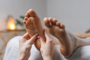 Eller nazikçe masaj masasındaki ayak tabanına masaj yapar. Yumuşak ışıklandırma ve doğal elementlerle çevrili rahatlatıcı bir spa ortamı vardır.